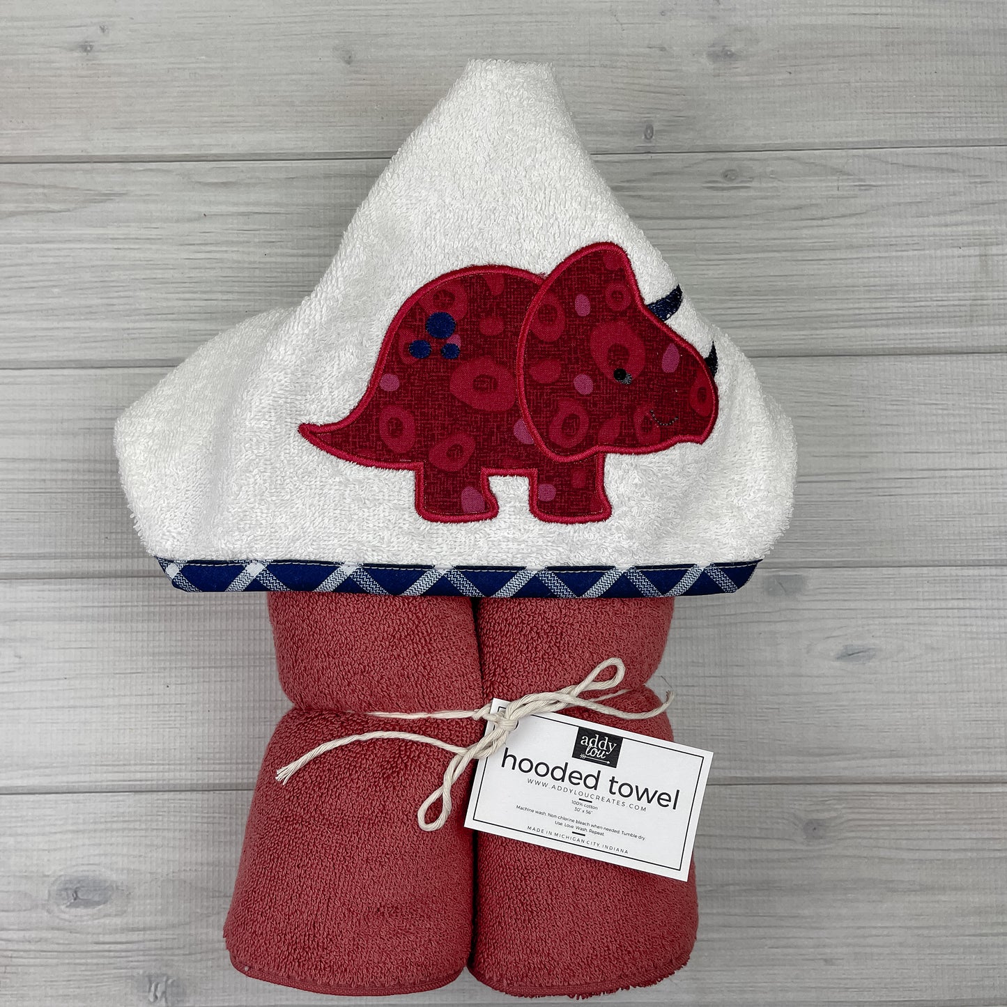 Dusty red hooded towel with red triceratops embroidered on a white hood. Binding is navy blue and white plaid