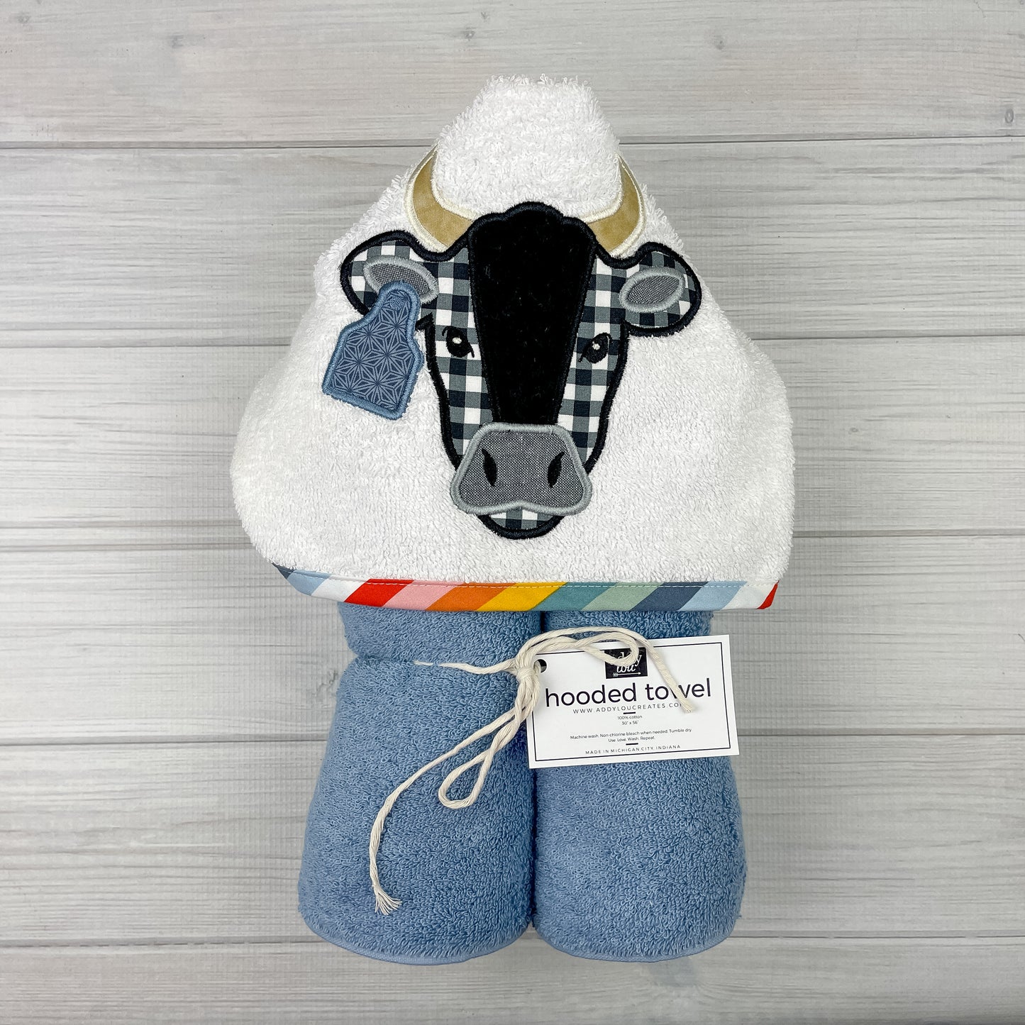 Medium blue hooded towel with black and white bull with horns and a tag embroidered on a white hood. Binding is muted rainbow colors of red, pink, orange, yellow, teal , pale green, dust blue, light blue and white.