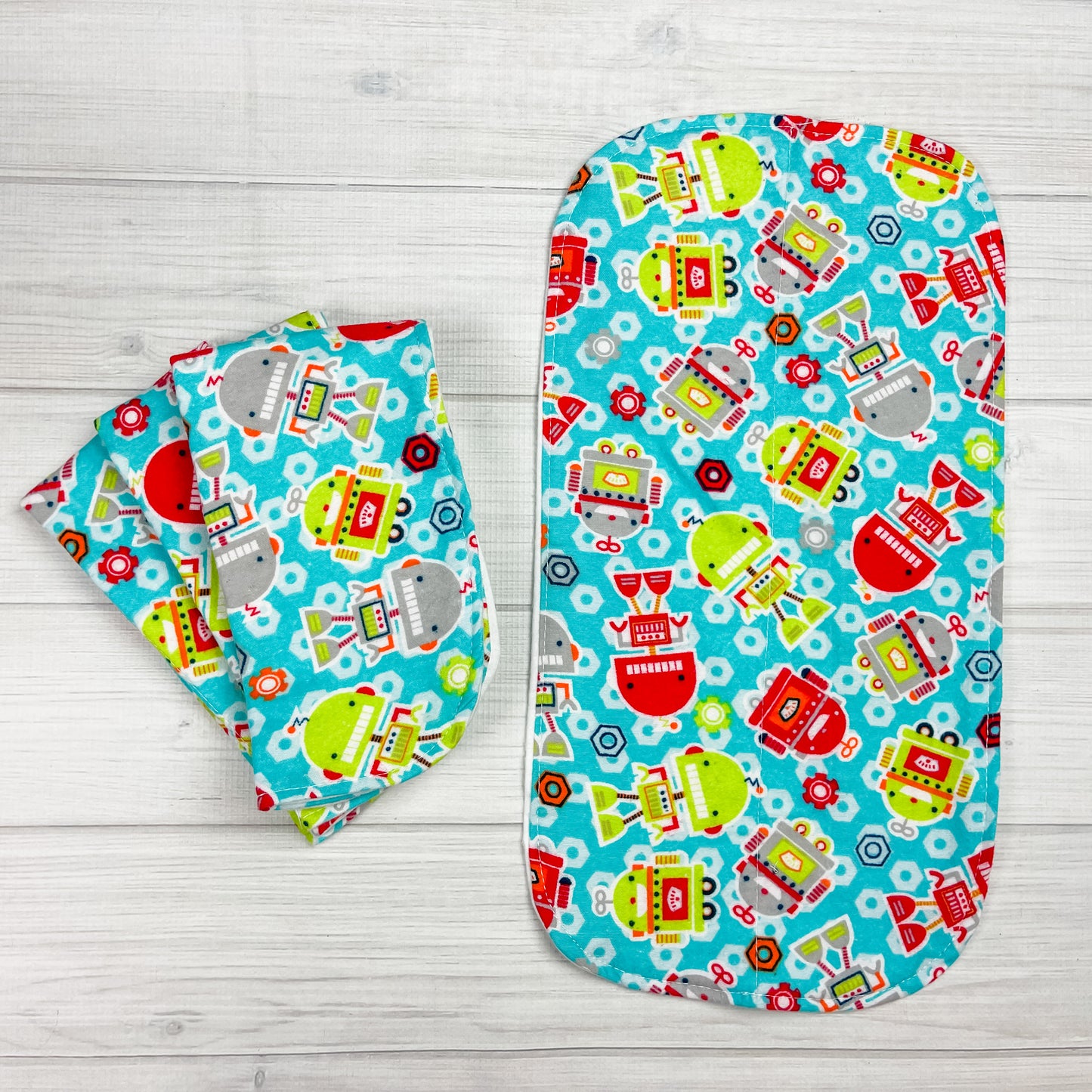 Flannel Burp cloths with robots in various sizes and shapes. Robots colors are in gray, lime green and red. The aqua blue background has nut in shades of red, orange and white.