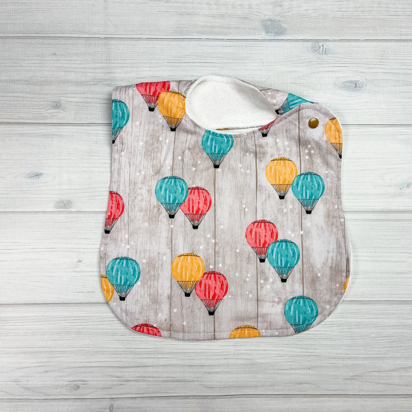 Generously sized infant/toddler bib with hot air ballons in teal, yellow and coral on a tan shiplapped backgrpund.